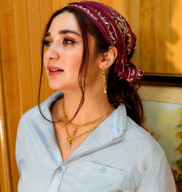 A woman wearing a burgundy paisley bandana headwrap, layered gold necklaces, and gold drop earrings, wearing a light blue button-up shirt against a warm wood-paneled background.